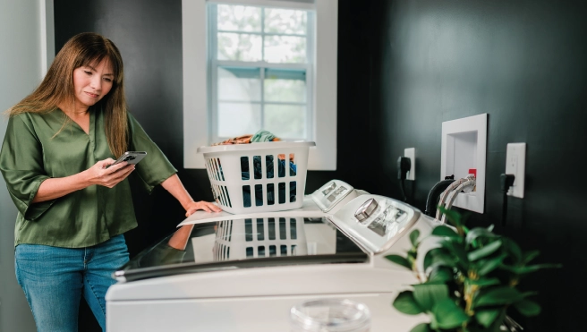 Women in green shirt and jeans looking at her phone while she stands next to her washing machine and dryer with a load of laundry.