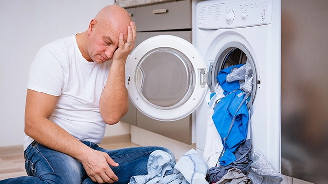 Tired man sits with laundry at the washing machine.