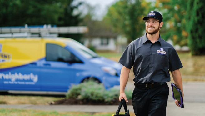 A professional from Dryer Vent Wizard walks up a driveway away from the company van to a house with toolbox and iPad.