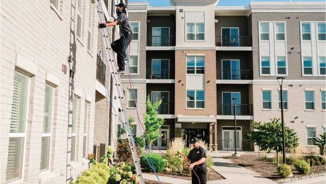 Dryer vent cleaning professional climbing ladder outside of apartment complex while the other stands below on the ground.