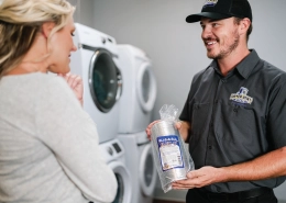 Woman and dryer vent professional speak while he holds a new dryer vent hose in front of stacked commercial dryers.