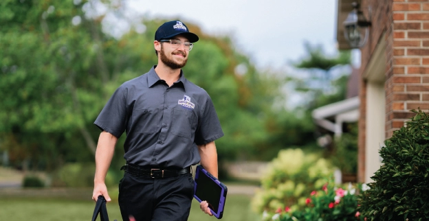 A uniformed dryer vent professional walks up to a house with tools smiling.