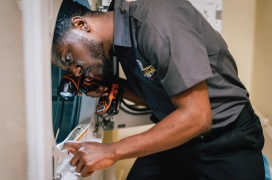 Male dryer vent cleaning professional peering into dryer while holding a tool.