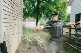 Male dryer vent cleaning professional stands outside of home holding cord while cleaning dryer vent.