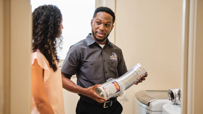 Male dryer vent cleaning professional holding part of a dryer vent duct while speaking to female homeowner.