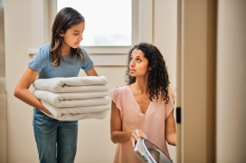 Mother kneeling and pulling clothes out of dryer while daughter holds folded towels.