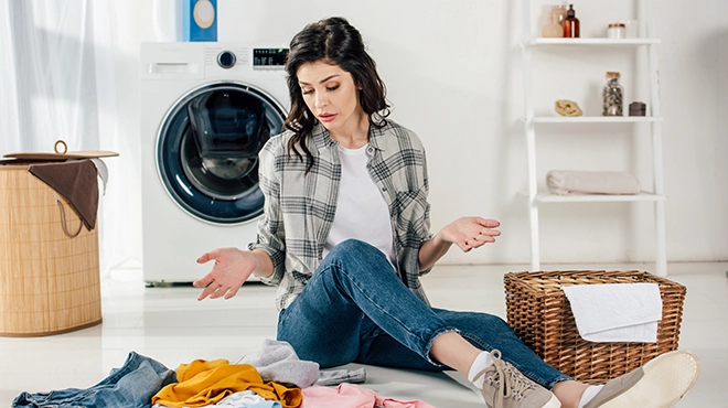 Confused woman sitting on floor near scattered clothes and baskets in laundry room.