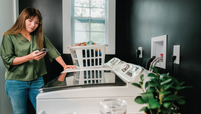 Female homeowner looks at cell phone while standing in tidy laundry room.