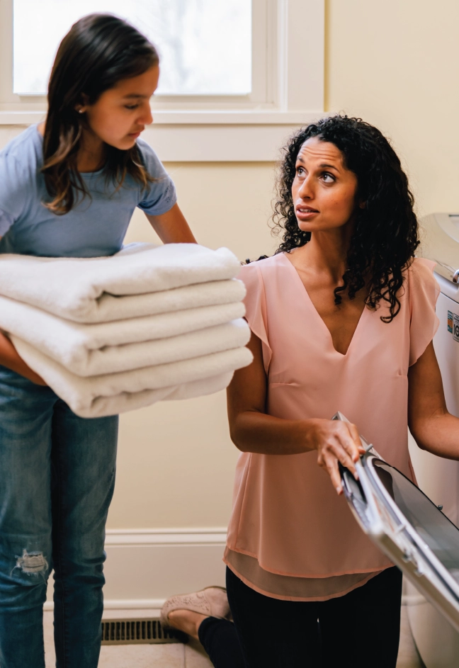 Mother kneeling and pulling clothes out of dryer while daughter holds folded towels.