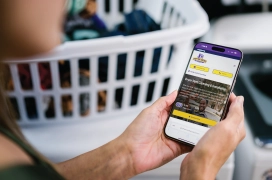 A woman looks at her cell phone and the Dryer Vent Wizard website for dryer vent cleaning services with a pile of laundry in front of her.