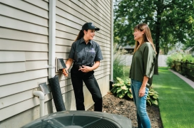 Female Dryer Vent Wizard employee takes a reading from the exterior of a home's dryer vent duct while talking with homeowner.